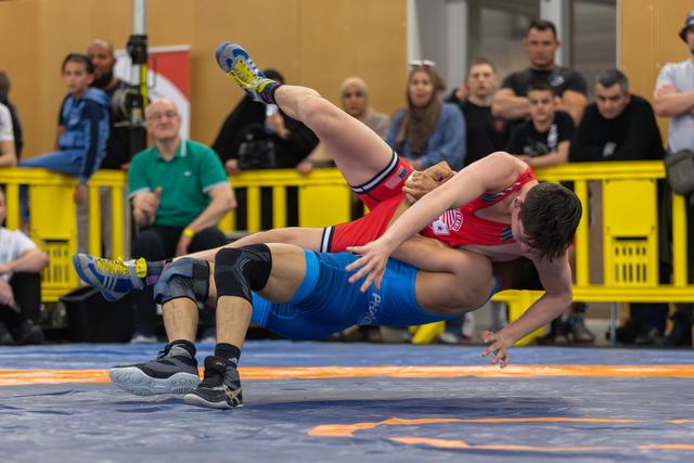 Wrestler in blue singlet executes a powerful throw, lifting red-clad opponent mid-air over the mat before spectators.