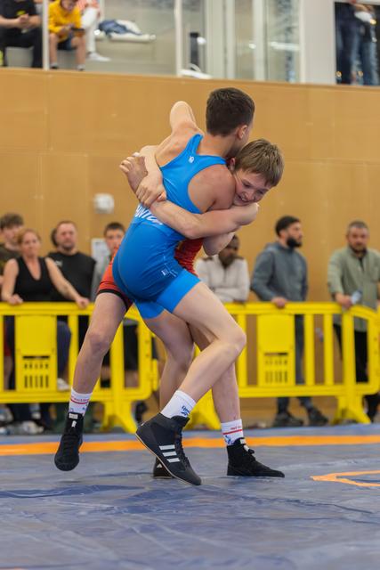 Wrestler in blue singlet lifts his smiling opponent off the mat during an intense grappling exchange indoors.