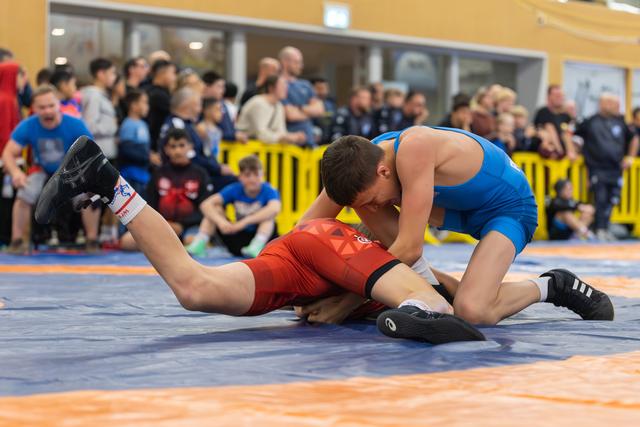 Wrestler in blue singlet dominates opponent in red, pressing him down on the mat while crowd watches intently.