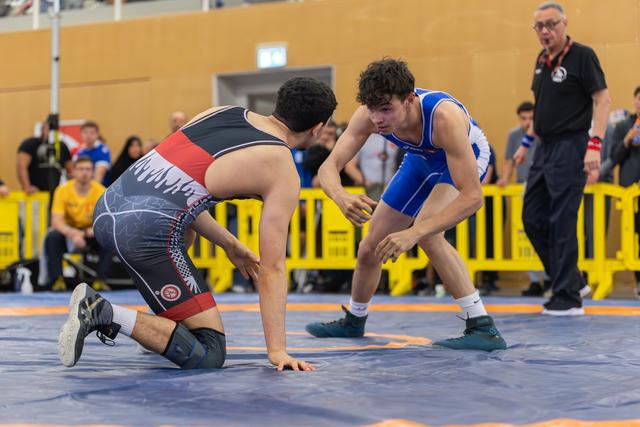 Young wrestler in blue singlet squares off aggressively over opponent on his knees on blue mat, referee watching.