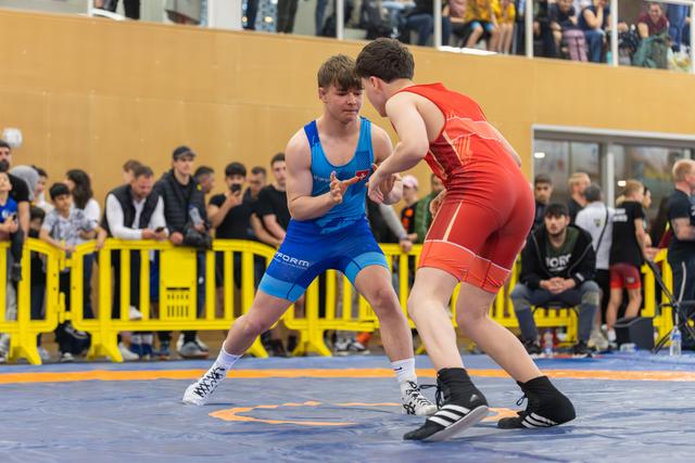 Two teenage wrestlers circle each other on the mat, focused and tense, as a crowd watches from behind yellow barriers.