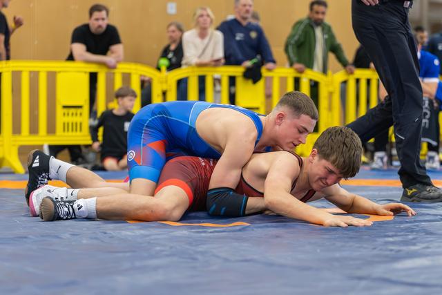 Young wrestler in blue singlet controls opponent on the mat, pressing him down with focused intensity during a match.