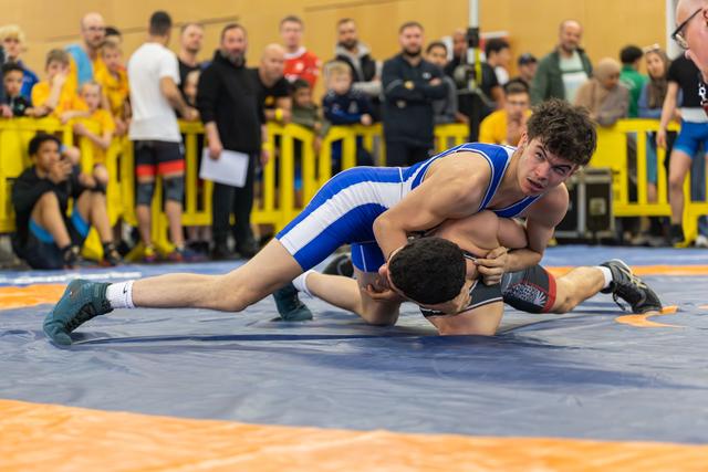 Young wrestler in blue singlet dominates opponent, pressing him to the mat with intensity before a watching crowd.