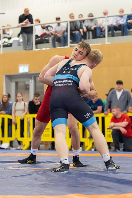Wrestler in black-blue Clausen NKC singlet locks a powerful clinch grip on red-suited opponent on mat.