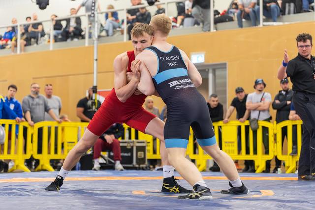 Wrestler in red and Clausen of NKC locked in intense grip battle on blue mat, crowd watching from above.