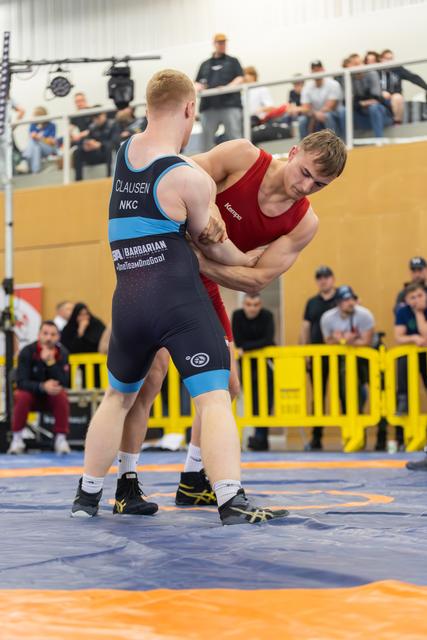 Wrestler Clausen in blue singlet locks arms with red-clad opponent in intense clinch on blue mat, crowd watching
