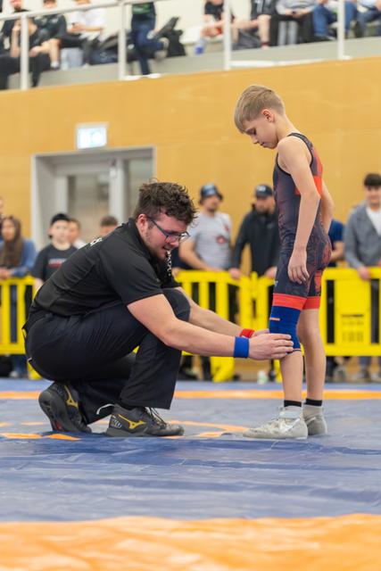A coach kneels to adjust a young boy's knee brace on the mat, the child looks down calmly before his match.