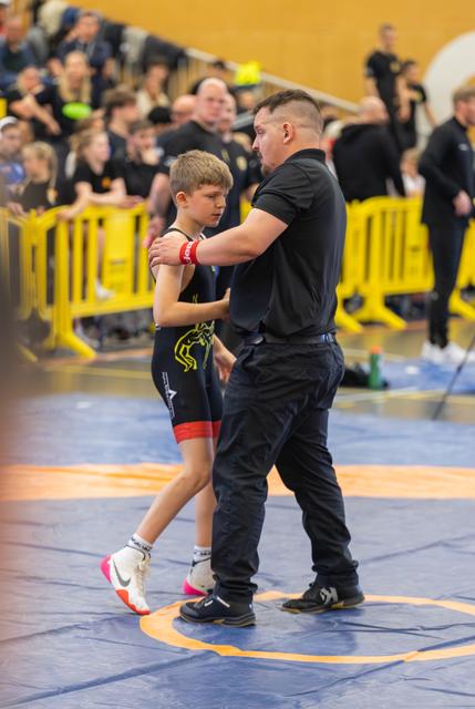 A coach steadies a focused young wrestler on the blue mat, offering guidance during a break in the bout, crowd watching behind.