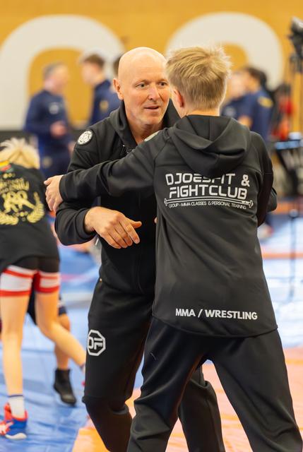 Bald coach embraces a young MMA wrestler in a Djoebfit Fight Club jacket, sharing a warm mentor moment on the mat.
