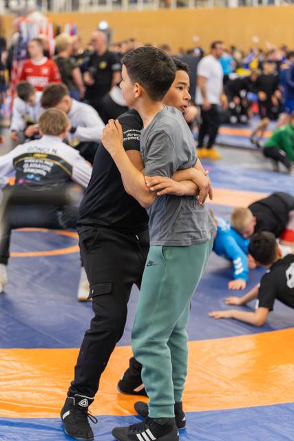 A coach in black holds a young athlete in green pants, demonstrating a wrestling grip on a busy blue mat.