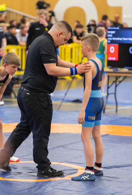 Coach places hand on young wrestler's shoulder, offering focused pre-match guidance on the blue mat during a youth event.