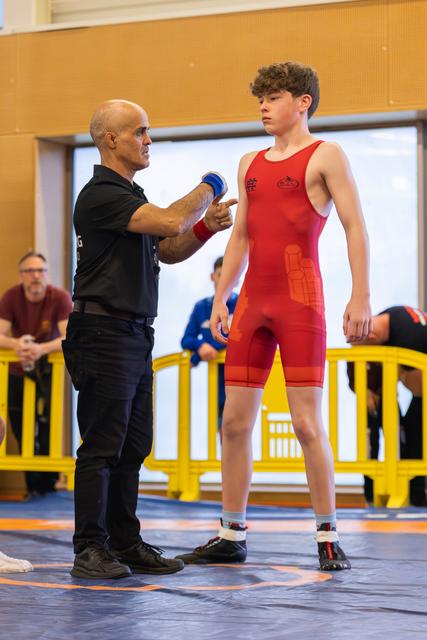 A bald coach points at a focused teenage wrestler in red singlet, offering pre-match guidance on the blue mat.