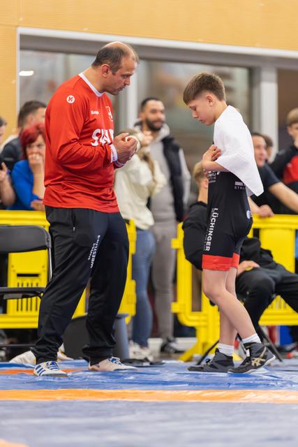 A coach in red reviews notes while a focused young wrestler listens intently, preparing between bouts on the mat.