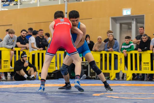 Wrestler Goodwin in red singlet locks up with blue-clad opponent on competition mat, crowd watching behind yellow barriers.