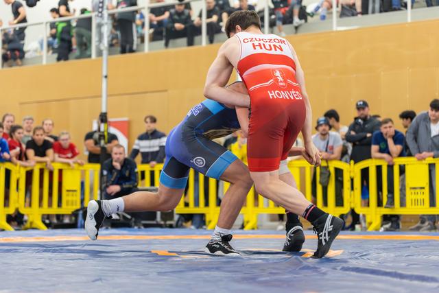 Czuczor (HUN/Honvéd) in red locks up with a blue-suited opponent on the mat, crowds watching behind barriers.