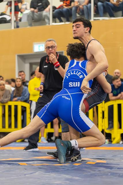 Swiss wrestler Jollien in blue SUI singlet grapples opponent on mat, referee watches intently in background.