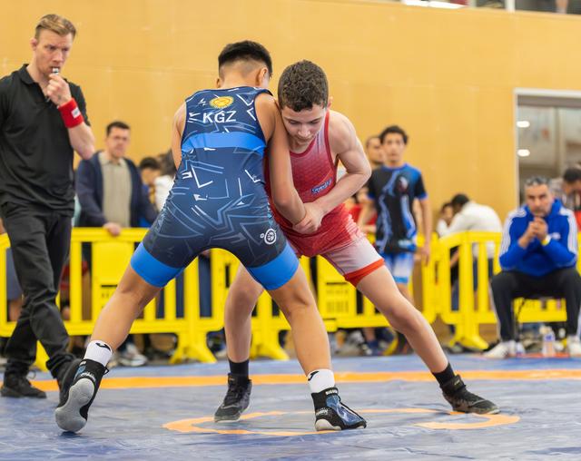 Young KGZ wrestler in blue singlet clinches with red-uniformed opponent on mat, referee watching closely behind.