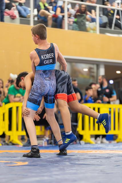Young wrestler Kober (GER) lifts his opponent off the mat during an intense youth wrestling bout before a crowd.