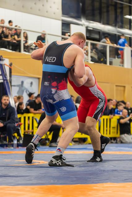 Dutch wrestler in black singlet locks up with red-suited opponent on orange mat, both straining forward under crowd's gaze.