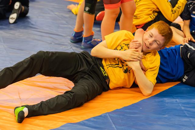 Laughing red-haired boy pinned on orange mat by opponent in blue shirt during youth wrestling match