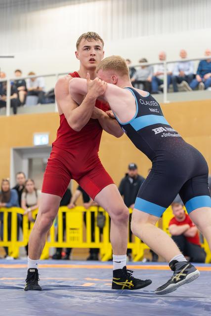 Young wrestler in red singlet controls opponent CLAUSSEN in blue, both locked in intense grip battle on blue mat.