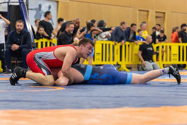 Wrestler in red singlet controls his opponent on the blue mat, leaning forward with focus during an indoor sports event.