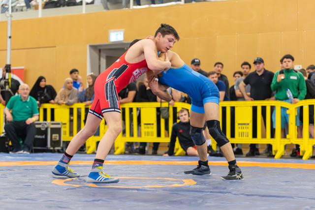 Two young wrestlers locked in an intense clinch on the blue mat, crowd watching behind yellow barriers.