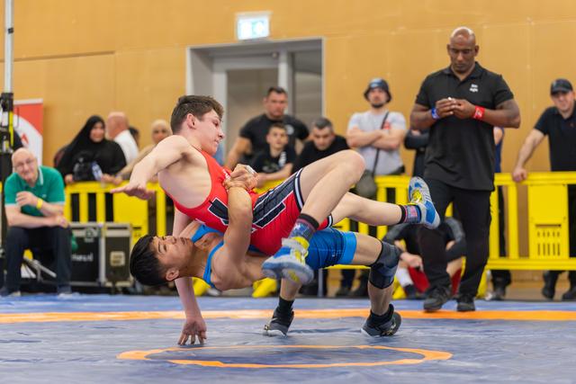 Wrestler in red lifts opponent in blue off the mat in a powerful throw, referees and crowd watching intently.