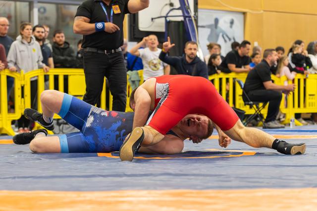 Wrestler in red singlet pins opponent in blue to the mat, crowd reacting with excitement in the background.