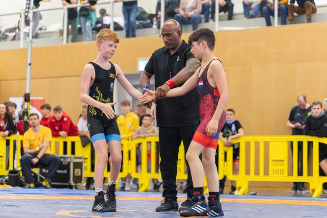 Referee joins hands of two young wrestlers in a pre-match handshake on the blue mat, crowd watching behind barriers.
