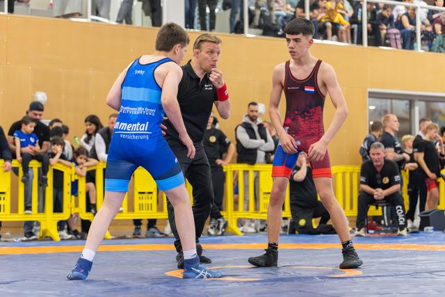 Referee gives instructions to two teen wrestlers—one in blue, one in dark red—before their bout on a blue mat.