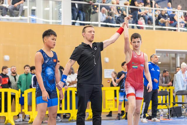 Referee raises the arm of a young wrestler in red, declaring victory, while the opponent in blue looks on stoically.