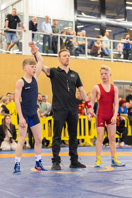 Referee raises the fist of a victorious boy in blue as the runner-up in red stands quietly beside them.
