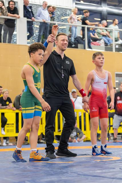 Smiling referee raises the hand of a young wrestler in green, declaring victory, as the opponent in red stands nearby.