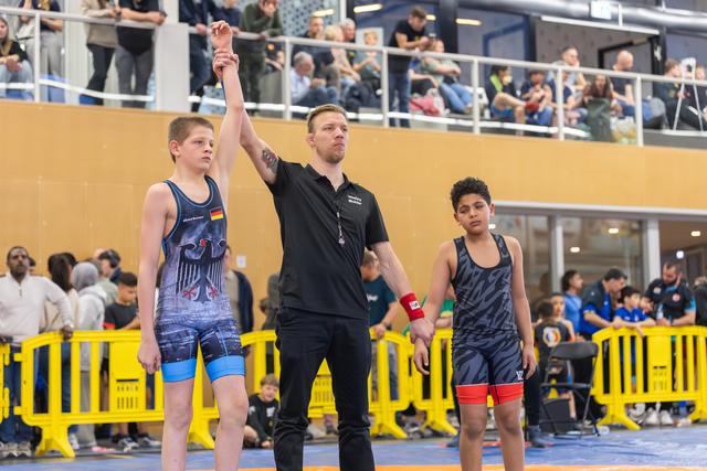 Referee raises the arm of a young German wrestler in victory, while his opponent stands nearby on the mat.
