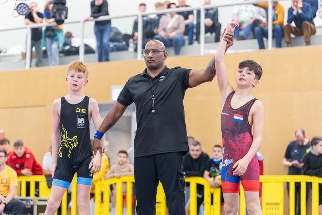 Referee raises the arm of a victorious boy in red, while the runner-up in black stands beside him, indoors.