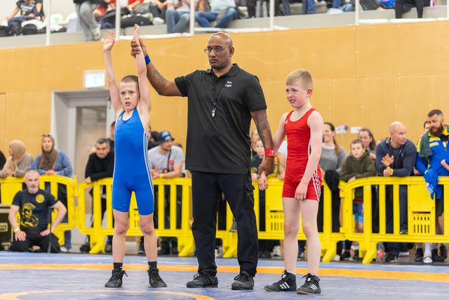 Referee raises the arm of a young boy in blue singlet declaring him winner, as defeated boy in red stands beside.