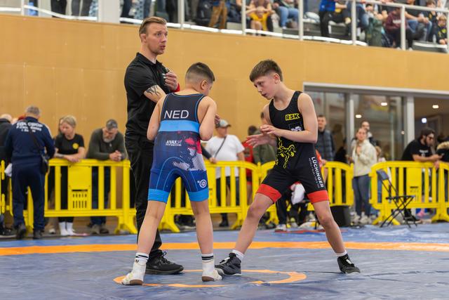 Young wrestler in NED singlet faces opponent in RSK kit as referee steps in to separate them on the blue mat.