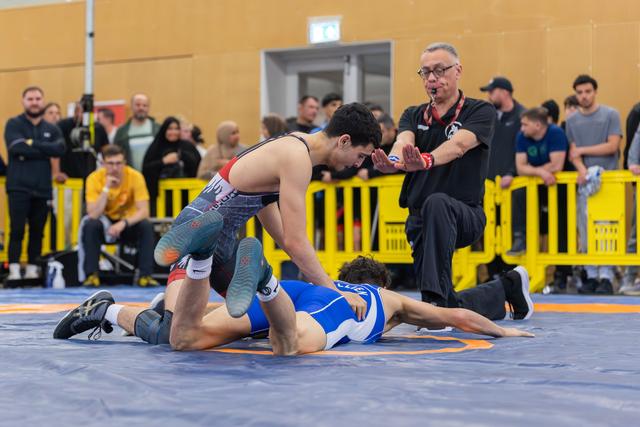 Wrestler in red pins opponent in blue to the mat as referee signals, crowd watching intently in background.
