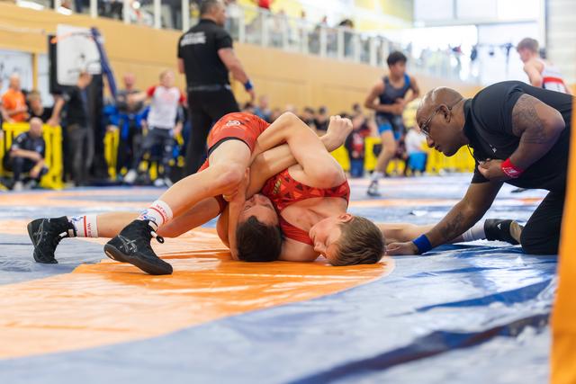 Two young wrestlers grapple on an orange mat as an intense coach leans in close, watching a pin attempt unfold.