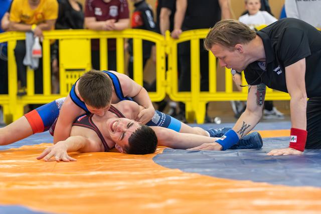 A wrestler pins his opponent to the orange mat, grinning, as a tattooed referee leans in closely to watch.
