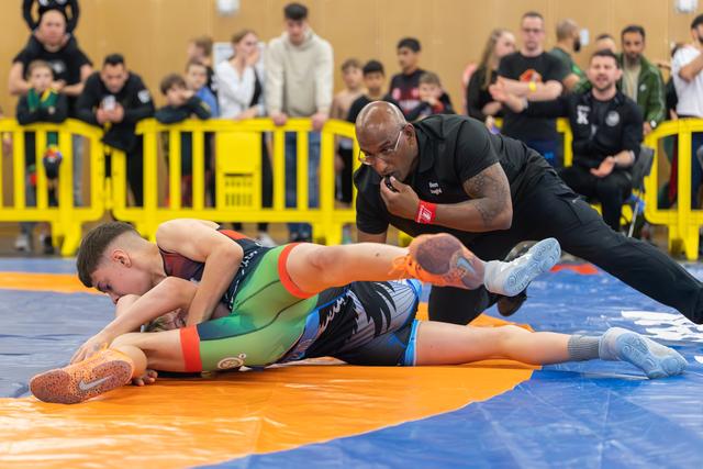 A referee leans in closely as one young wrestler pins another to the orange mat, crowd watching intently behind barriers.