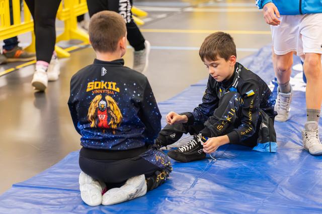 Two young Sokil Vinnytsia wrestlers sit on a blue mat, one carefully tying his shoes before the match.