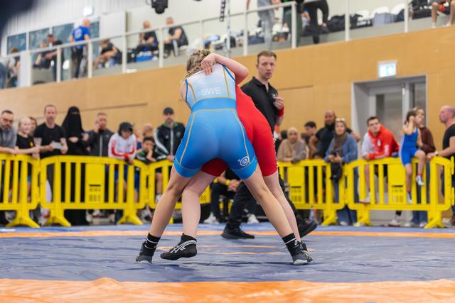 Swedish wrestler in blue singlet grapples opponent in red on mat, crowd watching from behind yellow barriers