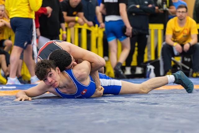 Young Swiss wrestler in blue singlet pinned on the mat, straining against opponent, crowd watching in background.