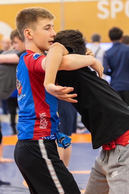 Young wrestler in blue-red jersey locks arms around opponent in black, intense focus during youth wrestling match