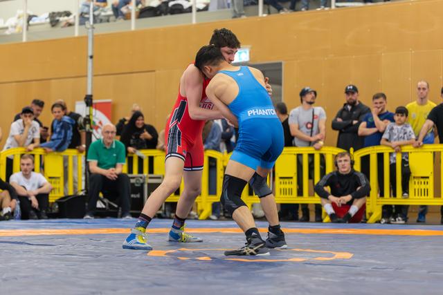 Two wrestlers lock in a tight clinch on the mat, both straining with intense focus during an indoor bout.