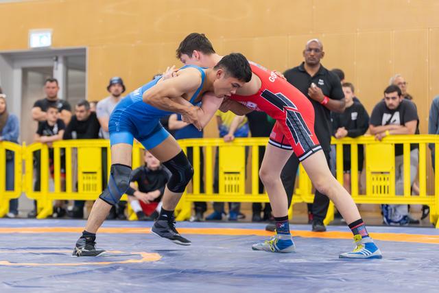 Two young wrestlers locked in intense grip, blue vs red singlet, leaning forward aggressively on competition mat with crowd watching.