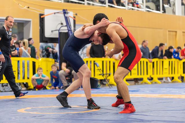 Two wrestlers locked in a tense clinch on a blue mat, both leaning forward aggressively during a competitive bout.