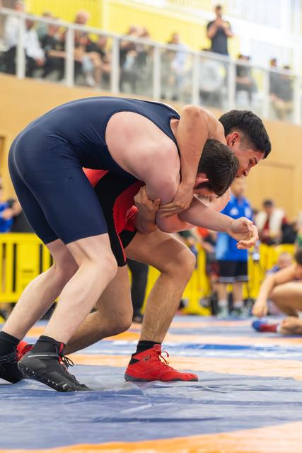 Two wrestlers locked in a tight clinch on the mat, one in navy blue singlet dominating over opponent in red, crowd watching.